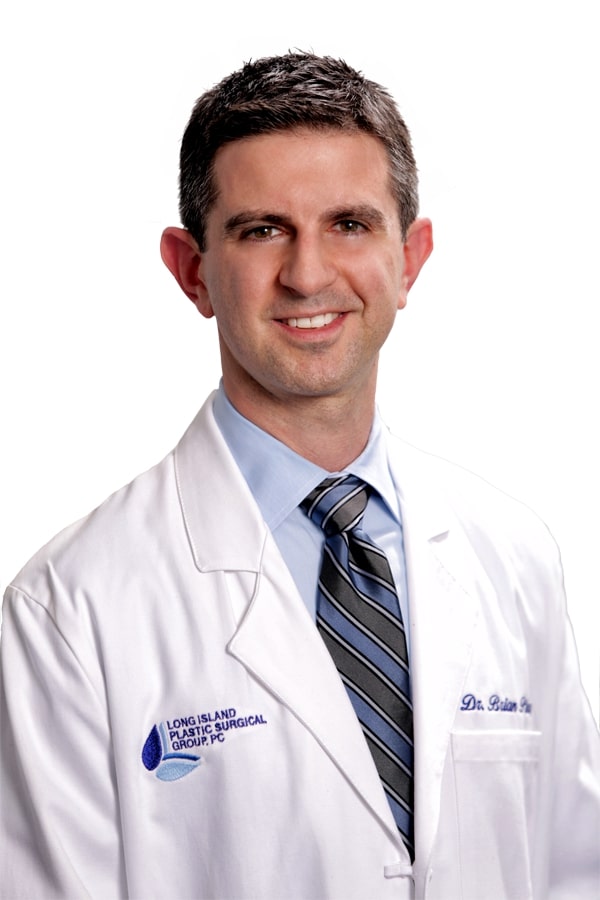 A man in a white medical coat and blue tie smiles at the camera. The coat has text that reads "Long Island Plastic Surgical Group" and a name embroidered.