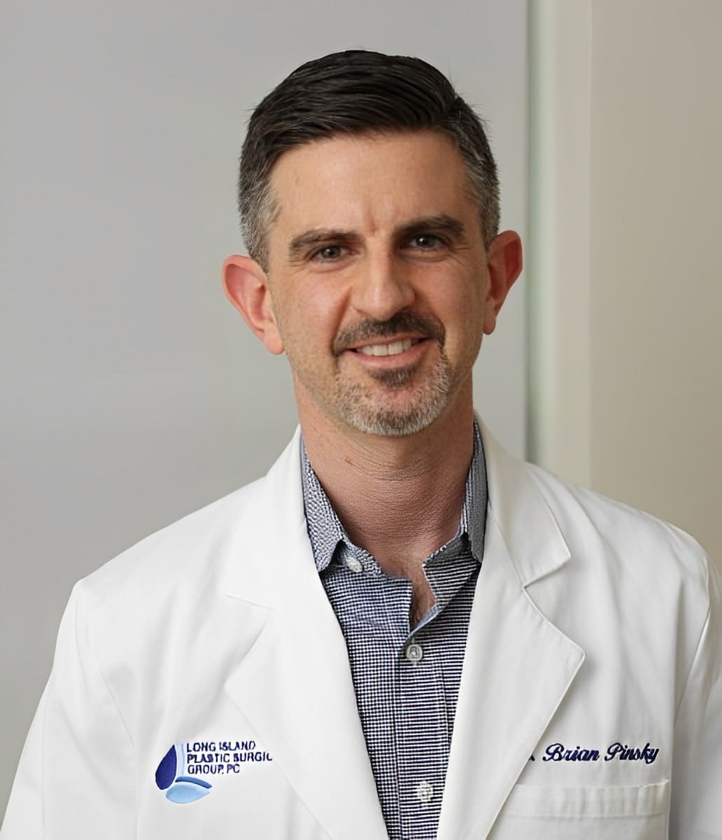 Man with short hair and facial hair wearing a white lab coat over a checkered shirt. The lab coat has text and a logo on it. He smiles softly while looking at the camera.