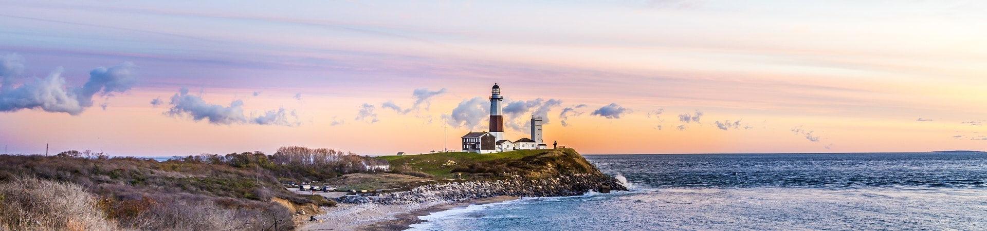 A lighthouse stands on a grassy hill by the ocean, with a rocky beach in the foreground. The sky is a blend of pastel colors during sunset.