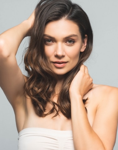 Woman with long brown hair poses with one arm raised, wearing a strapless top against a plain background.