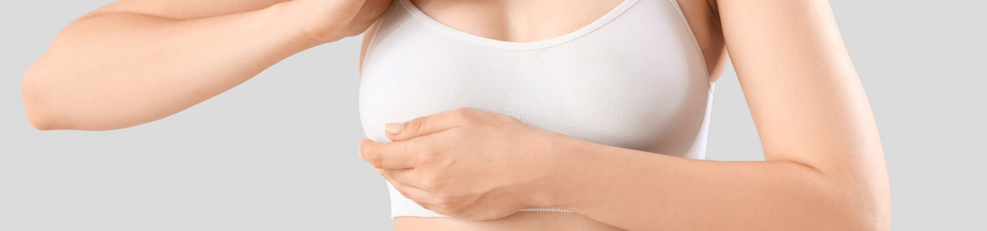 A person in a white tank top carefully examines their breast with one hand, contemplating the possibility of a breast lift, against a plain background reminiscent of Long Island's serene simplicity.