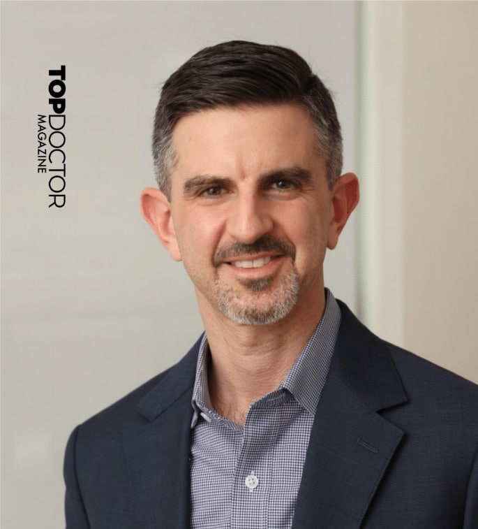 Man in a suit and collared shirt smiling with "Top Doctor Magazine" text beside him.