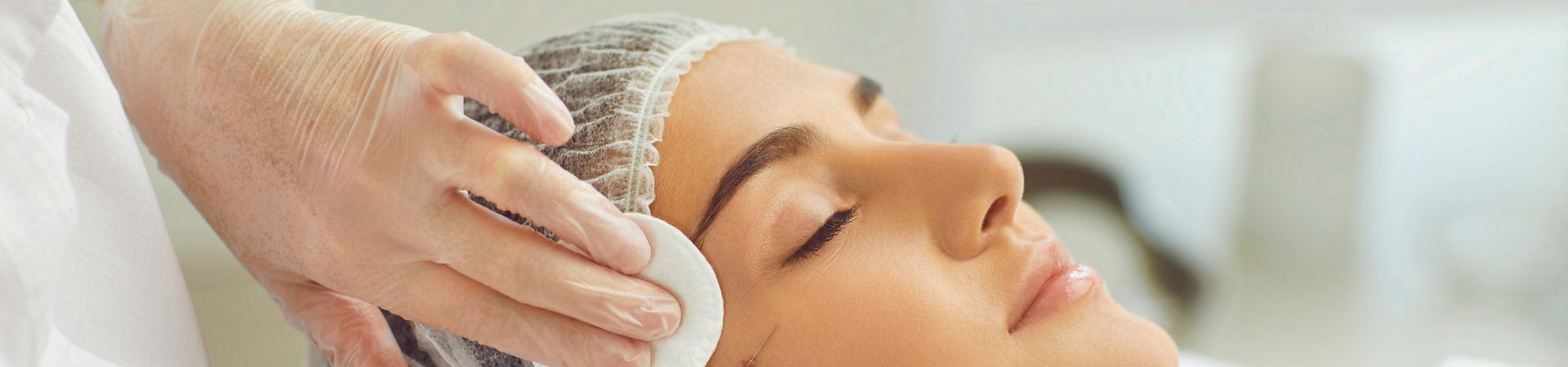 A person with a cap is lying down with eyes closed, while a gloved hand applies a cotton pad to their face, as part of a Botox treatment on Long Island.