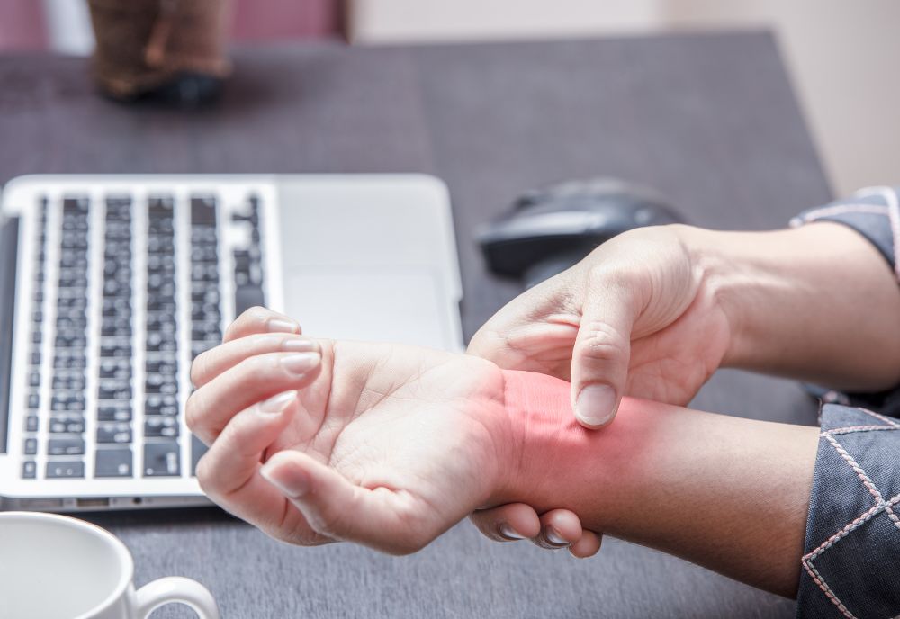 A person holding their wrist in pain, with a red area indicating discomfort. A laptop and mouse are visible on the table.