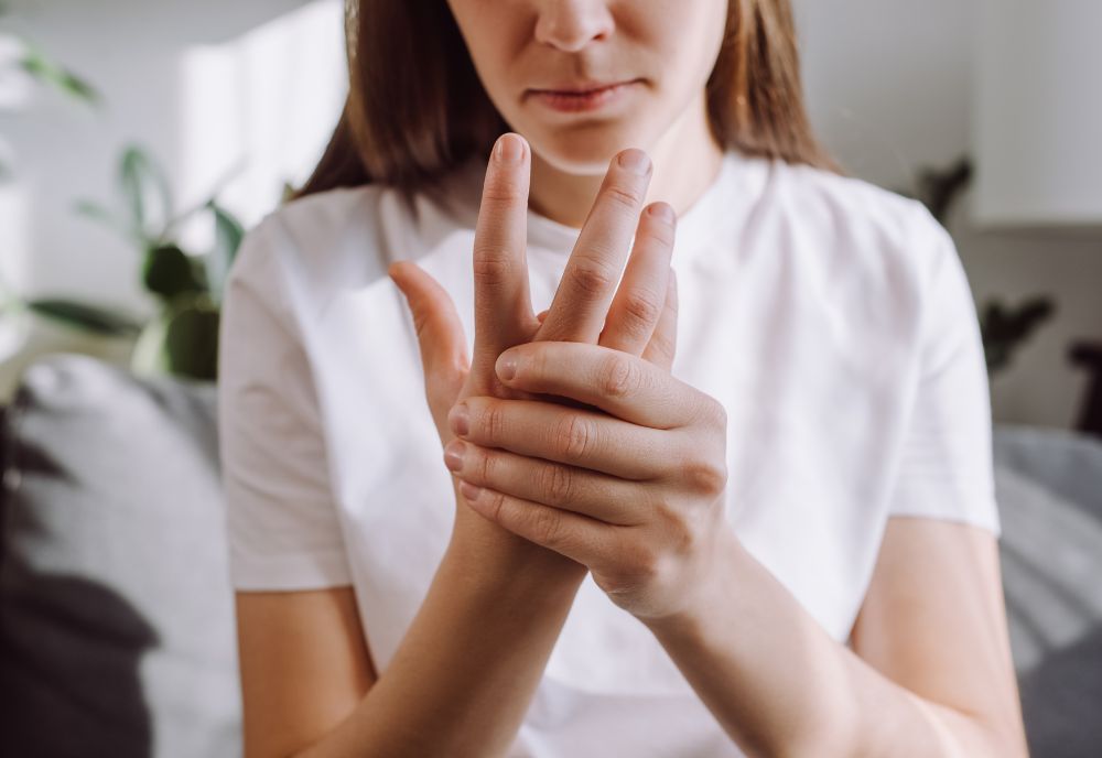 Person in a white shirt holding their hand, possibly indicating discomfort or self-care, with a blurred background of plants and a couch.