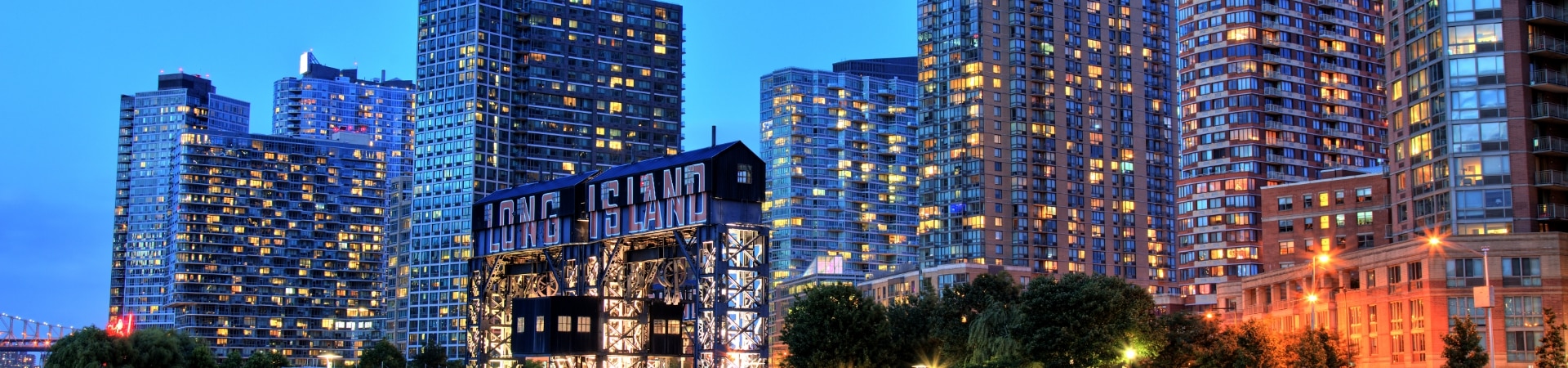 Cityscape at dusk showing high-rise buildings with illuminated windows and a Gantry Plaza State Park sign in the foreground.