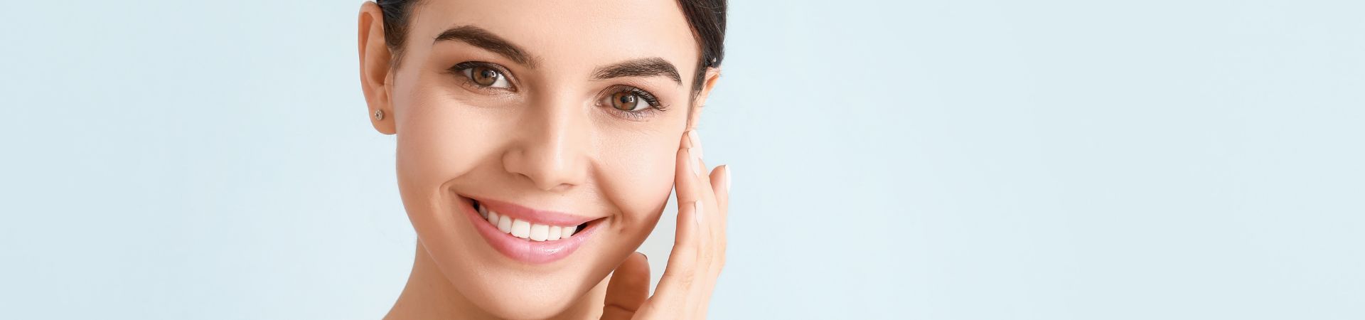 A person with dark hair smiles while touching their face against a light blue background, radiating the confidence that comes from a rejuvenating skin treatment on Long Island.