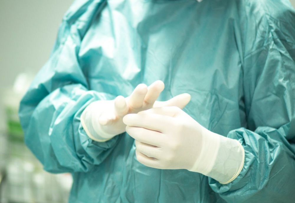 A person in a teal surgical gown is wearing white gloves in a medical setting.