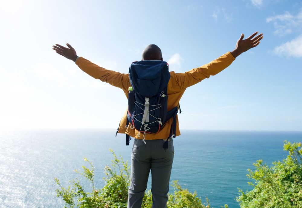 A person with a backpack stands on a cliff, arms outstretched, overlooking the ocean under a blue sky, capturing a scene worthy of Dr. Pinsky’s scenic inspirations.