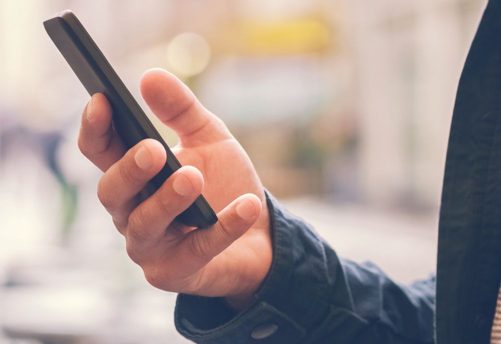 A person holds a smartphone in one hand. They are wearing a dark jacket. The background is blurred and outdoors.