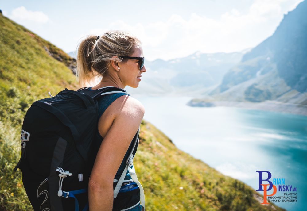 A person in sunglasses and a backpack stands on a grassy hillside, overlooking a lake and majestic mountains, enjoying the important tranquility that nature offers.