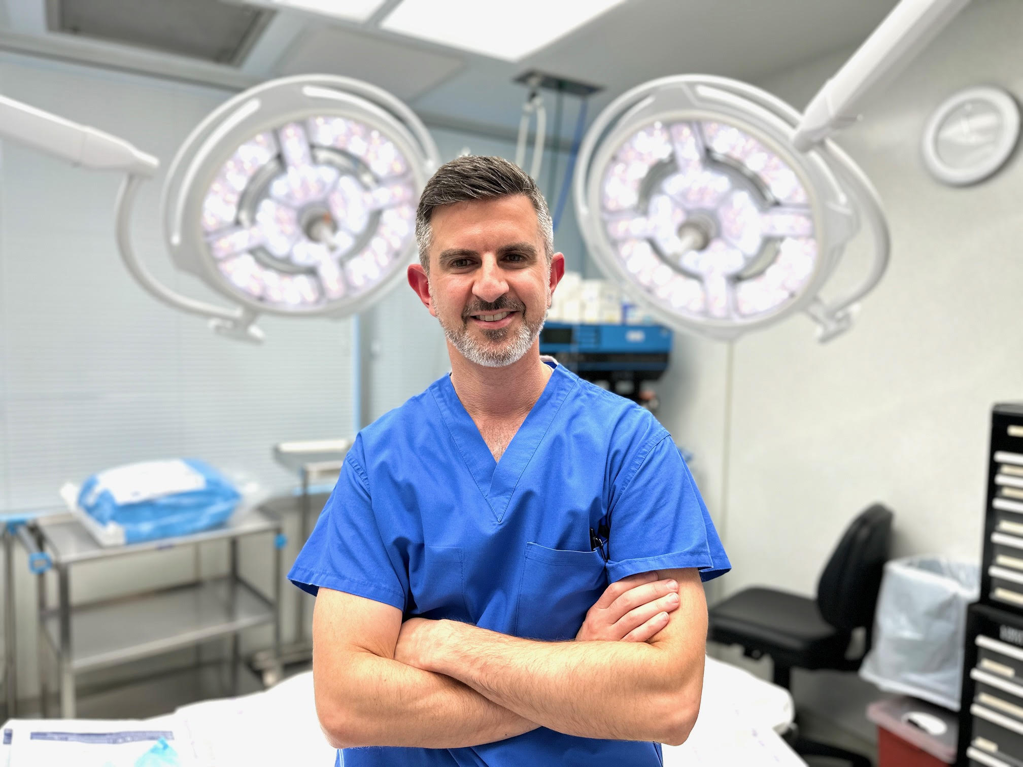 A man in blue scrubs stands with arms crossed in a brightly lit medical room, surgical lights and equipment in the background—an ideal image for a healthcare landing page.