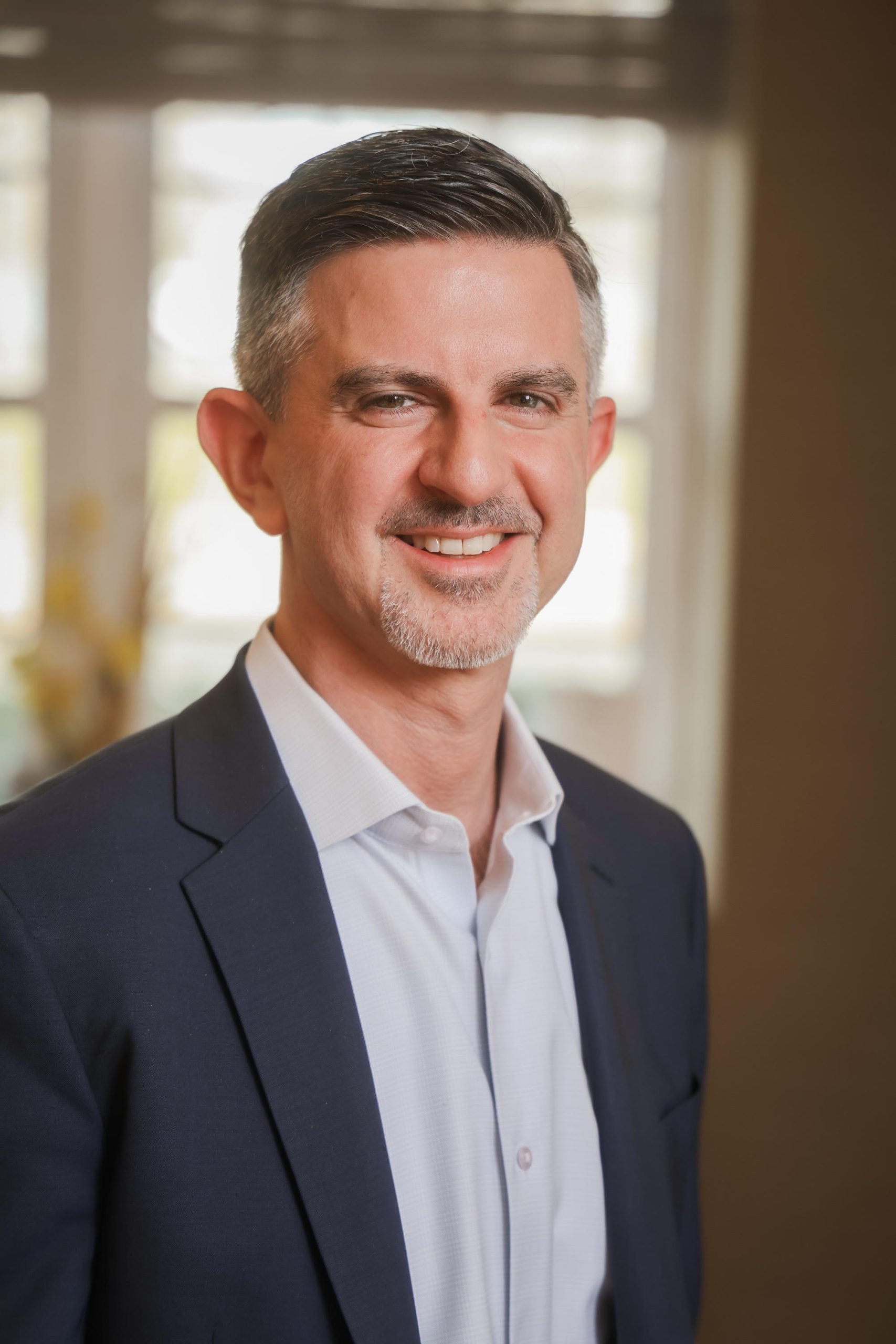 A man with short dark hair and a goatee, wearing a navy blazer and white shirt, smiles while standing indoors in front of a window—perfect for a professional landing page. A man with short dark hair and a goatee, wearing a navy blazer and white shirt, smiles while standing indoors in front of a window—perfect for a professional landing page