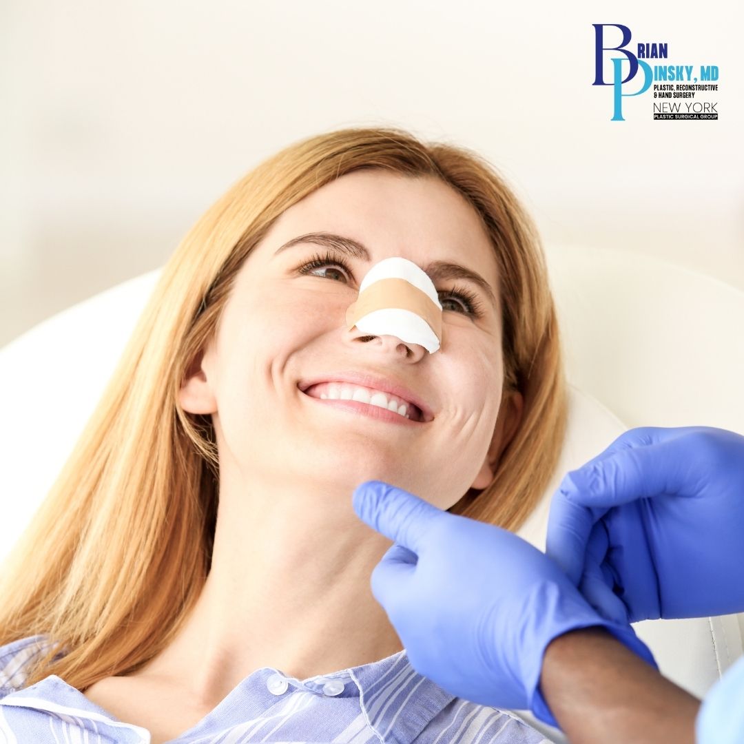 A woman with bandages on her nose smiles while a gloved medical professional examines her in a clinical setting after undergoing rhinoplasty on Long Island.