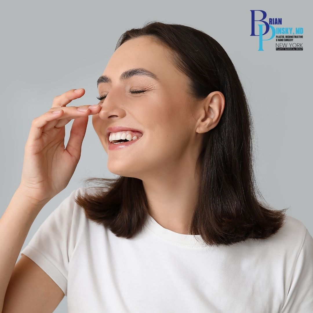 Woman with straight brown hair and a white shirt smiles with eyes closed, touching her nose with her right hand—an image reflecting confidence after rhinoplasty on Long Island. A medical clinic logo appears in the top right corner.
